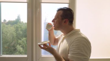 Genç ve yakışıklı bir adam pencereden dışarı bakan bir dairedeki fincandan kahve içer. Güzel bir gülümsemeyle sade kahve iç. Yavaş çekim.