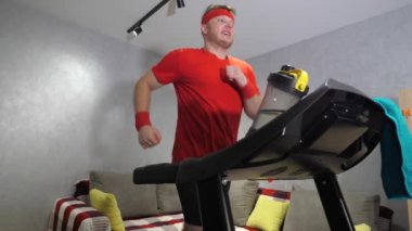Kırmızı spor üniformalı bir adam evdeki koşu bandında koşuyor. Kardiyo çalışması. Yavaş çekim.
