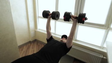 Spor bankında elinde dambıllar olan genç bir sporcu. Erkek göğüs kaslarının pompalanması. Evde idman yapıyorum. Yavaş çekim