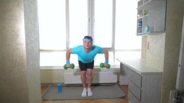 Ellerinde dambıllarla spor paspası üzerinde. Adam kol kaslarını şişiriyor. Ağır çekim. Evde egzersiz yap..