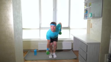 Ellerinde dambıllarla spor paspası üzerinde. Adam kollarının ve sırtının kaslarını pompalıyor. Ağır çekim. Evde egzersiz yap..