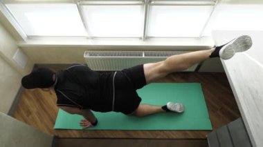 Genç bir atletik adam spor minderinde ısınıyor. Bacak esneme hareketi. Çömelirken bacaklarını vantilatörde salla. Ağır çekim. Evde egzersiz yap..
