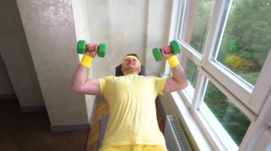 Dambılı komik bir sporcu. Evde antrenman yapan genç ve komik bir adam. Dambıllarla egzersiz. Yavaş çekim.