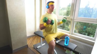Elinde dambıllar ve müzik kulaklıkları olan genç bir sporcu. Kol kaslarını şişirmek için dambıllarla evde egzersiz yapmak. Yavaş çekim
