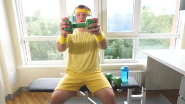 Dambılı komedyen kulaklıkla müzik dinlerken kas yapıyor. Evde antrenman yapan genç ve komik bir adam. Dambıllarla egzersiz. Yavaş çekim.