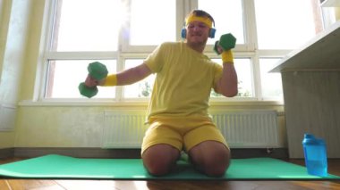 Elinde dambıllar olan bir sporcu kulaklıkla müzik dinlerken dans eder. Genç bir adam için ev antrenmanı. Spor minderinde neşeli ve pozitif bir sporcu. Yavaş çekim.