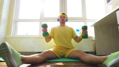 Elinde dambıllarla spor paspasıyla evde antrenman yapıyor, genç bir adam spor egzersizleri yapıyor. Yerde egzersiz yapan çekici, aktif bir adam. Yavaş çekim