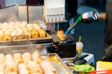 Satıcı peyniri eritmek için mutfak meşalesini kullanır, hafif kavrulana kadar mutfak meşalesinin alevi üzerinde hafifçe çalışır.