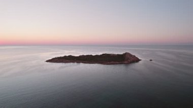 Aerial view of island in Mediterranean sea, near Sardinia during sunrise. Drone orbit shot. Calm sea with a little ripples. Clear blue sky with red Sun raising up.
