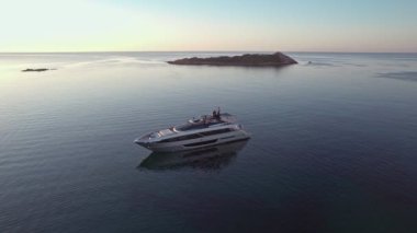 Aerial orbit view of a wealthy billionaire luxury Riva Corsaro super motor yacht staying at anchor in mediterranean sea during colorful sunrise with green islands in the beautiful bay