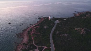 Deniz kenarındaki deniz fenerine giden ve çatıda yanıp sönen bir ışıkla çekilmiş bir hava görüntüsü. Etrafı yeşil tepeler ve ağaçlarla çevrili