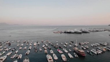 Napoli, İtalya 'da bir marinanın havadan görünüşü. Sakin sularda demirli tekneler ve lüks yatlar. Gün batımında yumuşak bir ışık, ufukta dağlar ve adalar, barışçıl Akdeniz atmosferi yaratıyor..
