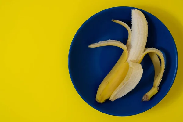 Half peeled banana on dark blue plate, both on yellow background. Minimalist style photography. Color theory. Complementary colors
