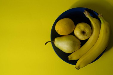 Yellow banana, apple, pear and lemon on blue plate. Top view. Flat lay. Yellow background. Copy space. Negative space. Complementary color harmony. 