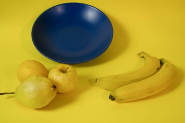 Blue plate on a yellow background with fruits around. Yellow banana, apple, pear and lemon. Top view. Flat lay. Yellow background. Copy space. Negative space. Complementary color harmony. 