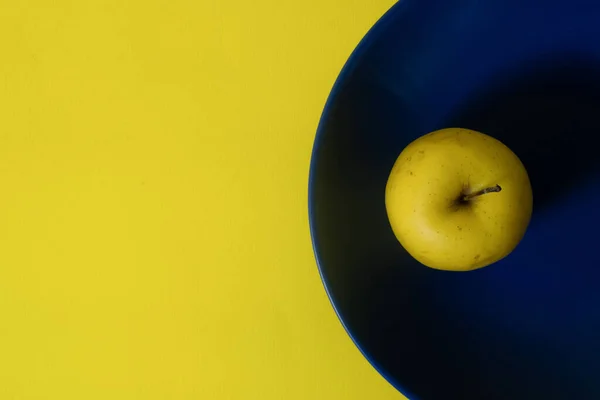 Top view of fresh yellow apple on a blue plate, both on yellow background, with copy space. Minimalist style photography. Yellow and blue color harmony. Color theory. Complementary color harmony. Top