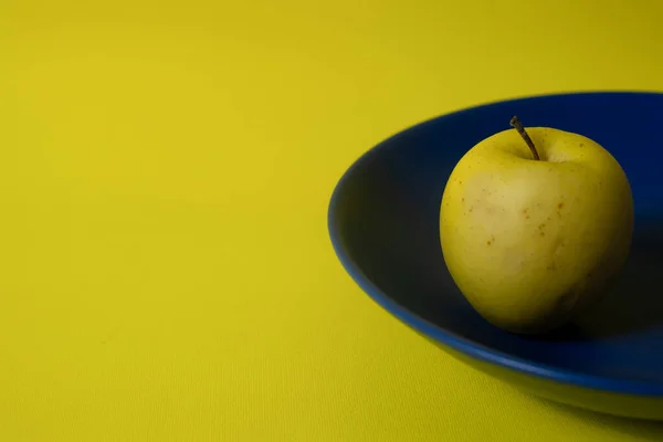 Fresh yellow apple on a blue plate, both on yellow background, with copy space. Minimalist style photography. Yellow and blue color harmony. Color theory. Complementary color harmony.