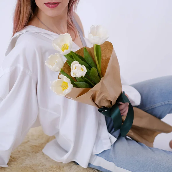 Woman in white shirt sitting and holding a bouquet of white tulips in her hands. Feminine. White shirt. Gift concept for woman, love, birthday, 8 March, mother day. Copy space.