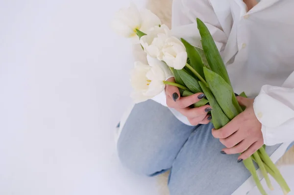 Woman in white shirt sitting and holding a bouquet of white tulips on a white background. Feminine. White shirt. Gift concept for woman, love, birthday, 8 March, mother day. Copy space.
