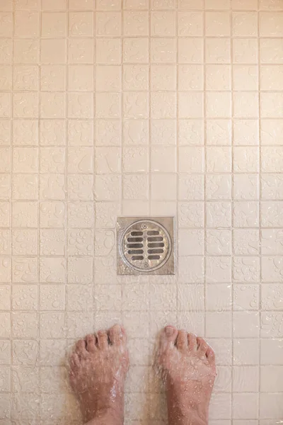 Bronzlaşmış insan ayakları üzerinde duştaki küçük kare bej fayansların üzerinde buhar ve suyla su sıçrıyor ve lağıma akıyor.