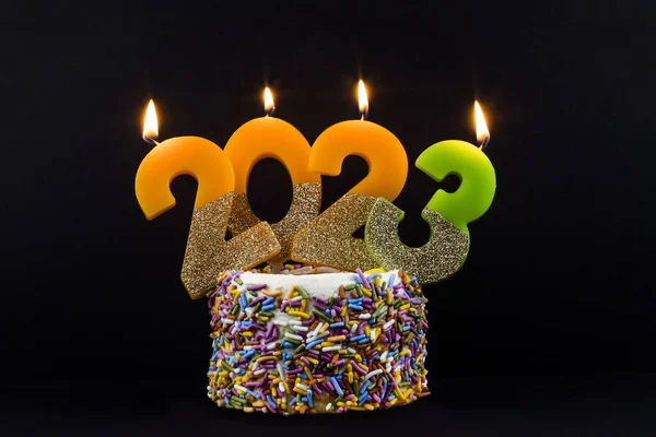 Closeup view of burning candles, making number 2023, on top of a cake with multi colored sprinkles, on black background. Happy New 2023 Year