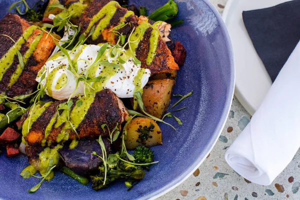 Closeup angle view of blackened salmon pieces, eggs, and mixed seasoned vegetables on the blue plate with a rolled white napkin