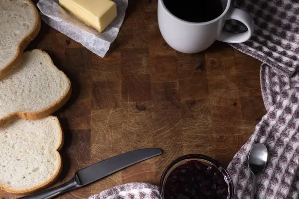 Closeup top angle view of breakfast items framinf a copy space. Pieces of bread, butter, jam, tea cup, kitchen towel and metal utencils around scratched wooden surface.