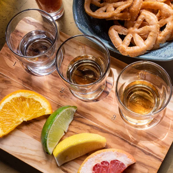 Tahta bir tahtanın üzerinde tekila, yanında meyve dilimleri ve atıştırmalık bir tabakla üç bardağın üst açı görüntüsünü kapatın. Tekila shot uçuşu. Kare resim