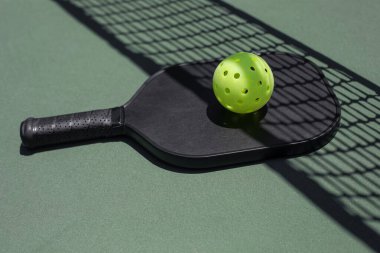 Closeup angle view of a black pickleball paddle with a yellow ball on top, laying on the court, the net shadow over it. clipart