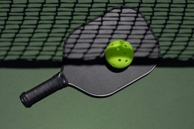 Üzerinde sarı bir top olan siyah bir picleball raketinin üst görüntüsü, yeşil kort yüzeyinde, üzerinde ağ gölgesi olan