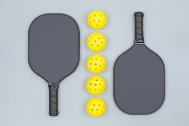 Açık gri arkaplan üzerinde beş sarı top bulunan iki siyah picleball raketinin düz görünümü