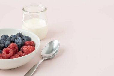 Böğürtlenli beyaz bir kase, bir bardak süt ve açık pembe arka planda fotokopi alanı olan bir kaşık.