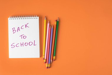 Okul tabelasına geri dönen beyaz spiralli defter ve turuncu arka planda fotokopi alanı olan renkli kalemlerin yukarıdan görünümünü kapat