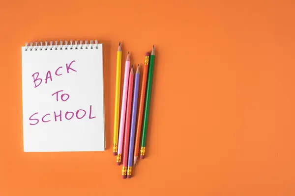 Okul tabelasına geri dönen beyaz spiralli defter ve turuncu arka planda fotokopi alanı olan renkli kalemlerin yukarıdan görünümünü kapat