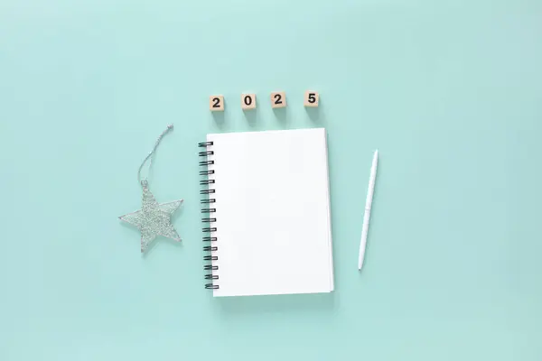 Açık beyaz not defteri, kalemi, gümüş yıldız süslemesi ve nane mavisi arkaplan üzerinde açık mavi metin için alanı olan 2025 numaralı düz çizgi. Yeni yıl planlama 2025