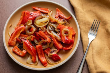 Sarı seramik tabağın üst açısı Shakshuka dolma biberli ve keten peçeteli soğanlı ve yan tarafında metal çatallı.