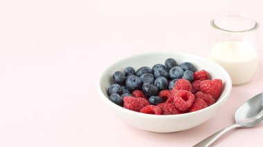 Beyaz bir kasede ahududu ve yaban mersinlerinin yakın görüntüsü, metal kaşık ve yanında küçük bir bardak süt, açık pembe arka planda yazı, afiş için boşluk