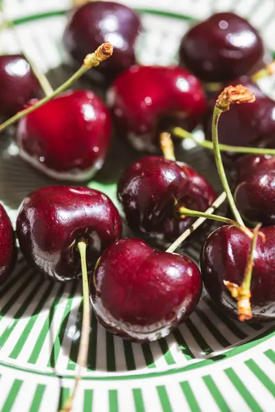 Dekoratif yeşil çizgili bir tabakta saplı taze koyu kırmızı kirazlar yakın plan, canlı bir detay ve yaz tazeliği ile doğal güneş ışığı altında yakalanır..