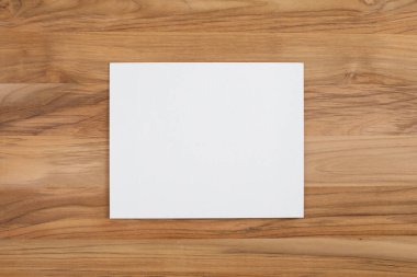 Doğal ahşap bir yüzeye yerleştirilmiş boş kare beyaz bir kartın yukarıdan aşağı görüntüsü. Markalaşma, davetiyeler, duyurular veya yaratıcı tasarım tasarımları için en düşük model arka plan.