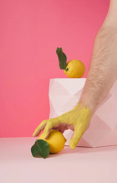 一只涂着黄色油漆的彩色手 正伸手去拿一个带有叶子的柠檬 放在粉红的背景上 该设计的特点是垂直框架 — 图库照片