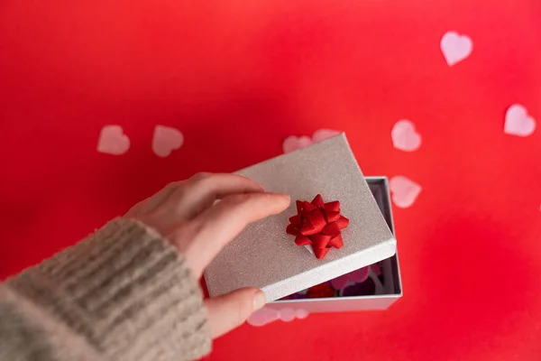 A womans hand opens a gift box with hearts on a red background.