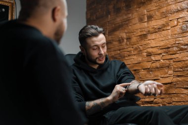 Dövme stüdyosunun içinde bir adam dövmelerini usta bir dövmeciye gösteriyor.