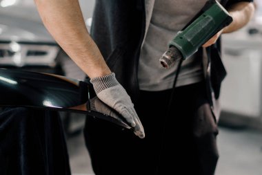 Bir işçi, atölyede saç kurutma makinesi ve kapanış spatulayla araba temizliği ve bakımı ile bir film ile bir araba spoiler kaplar