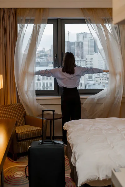 stock image young woman opening curtain while standing luxury hotel room looking through window enjoying city skyscrapers close up