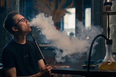 Gözlüklü bir adam geleneksel nargile piposu tüttürür. Bir adam nargile kafeteryasında ya da bar barında yoğun duman çıkarır.
