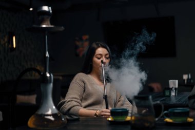 Güzel genç bir kadın nargile soluyor nargile içiyor bar konseptinde oturuyor ve sigara içiyor.