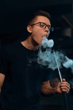 Gözlüklü bir adam geleneksel nargile piposu tüttürür. Bir adam nargile kafesinde ya da dinlenme odasında kalın dumanlı yüzükler solur.