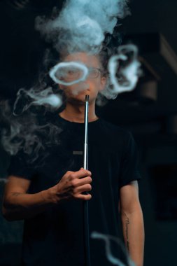 Gözlüklü bir adam geleneksel nargile piposu tüttürür. Bir adam nargile kafesinde ya da dinlenme odasında kalın dumanlı yüzükler solur.