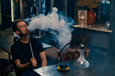 Gözlüklü bir adam geleneksel nargile piposu tüttürür. Bir adam nargile kafeteryasında ya da bar barında yoğun duman çıkarır.