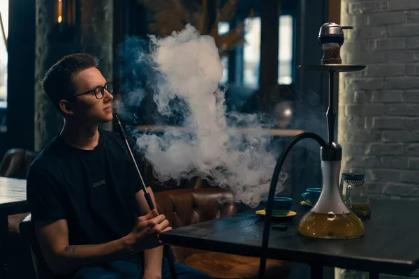 A man in glasses smokes a traditional hookah pipe A man exhales thick smoke in hookah cafe or lounge bar
